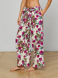 A person stands barefoot against a plain white background, wearing the Geraldine Cover-Up Pant—wide-leg linen resort pants featuring pink and purple flowers with green leaves and a comfortable tie waist.
