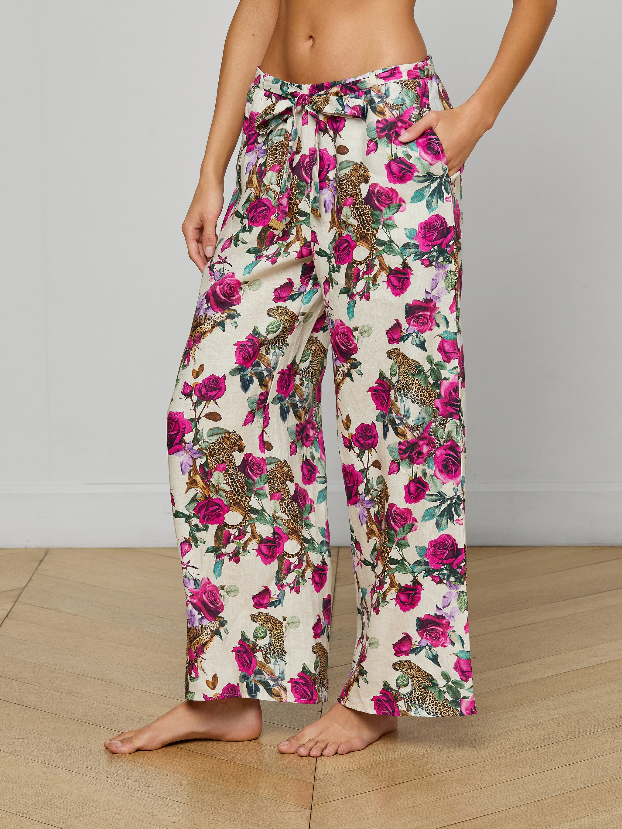 A person stands barefoot against a plain white background, wearing the Geraldine Cover-Up Pant—wide-leg linen resort pants featuring pink and purple flowers with green leaves and a comfortable tie waist.