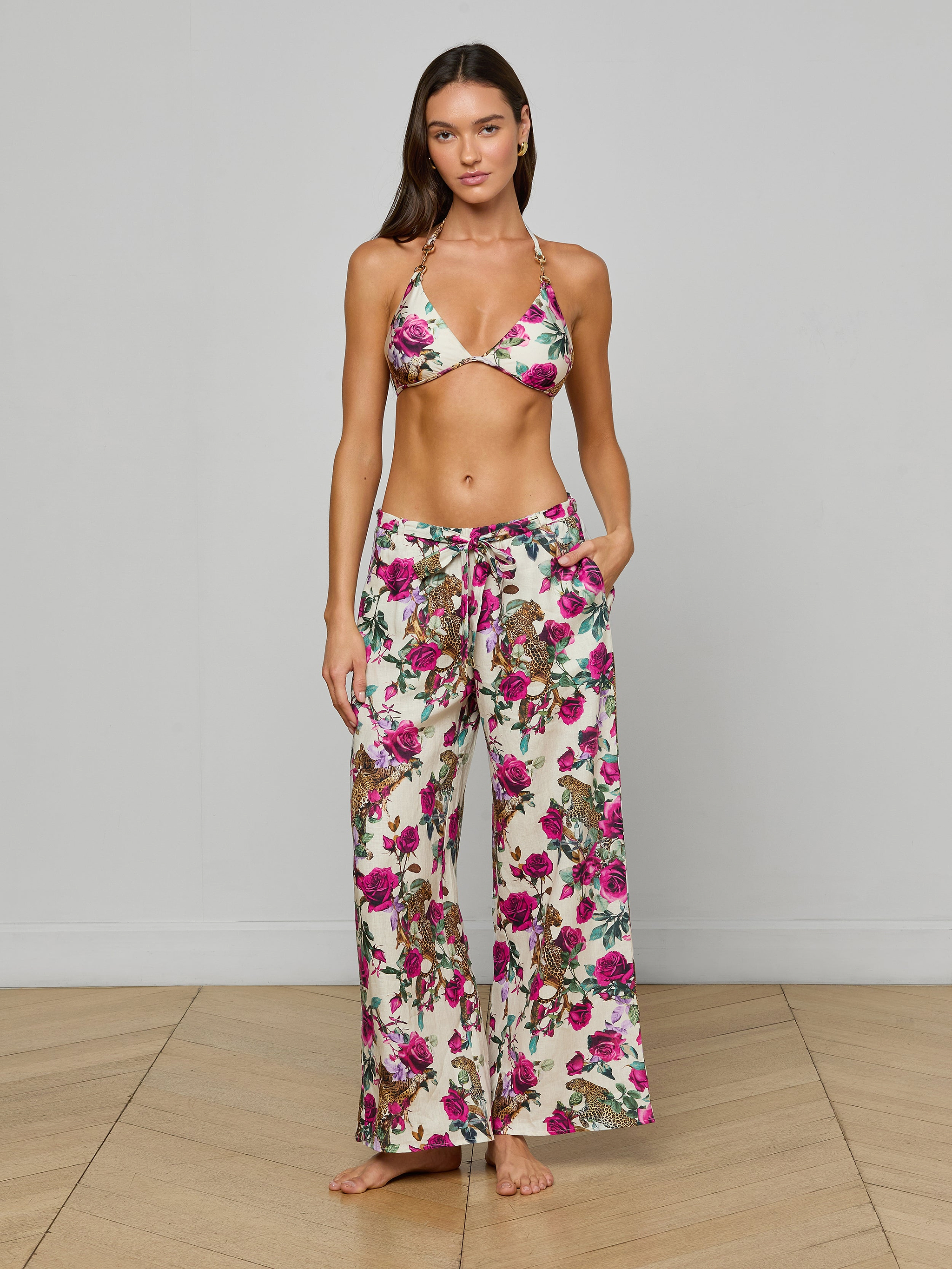 A woman stands barefoot against a plain light background, her hands tucked in her pockets, wearing the Geraldine Cover-Up Pant—a pair of loose linen resort pants with a tie waist in a rose leopard print—paired with a matching floral bikini top.