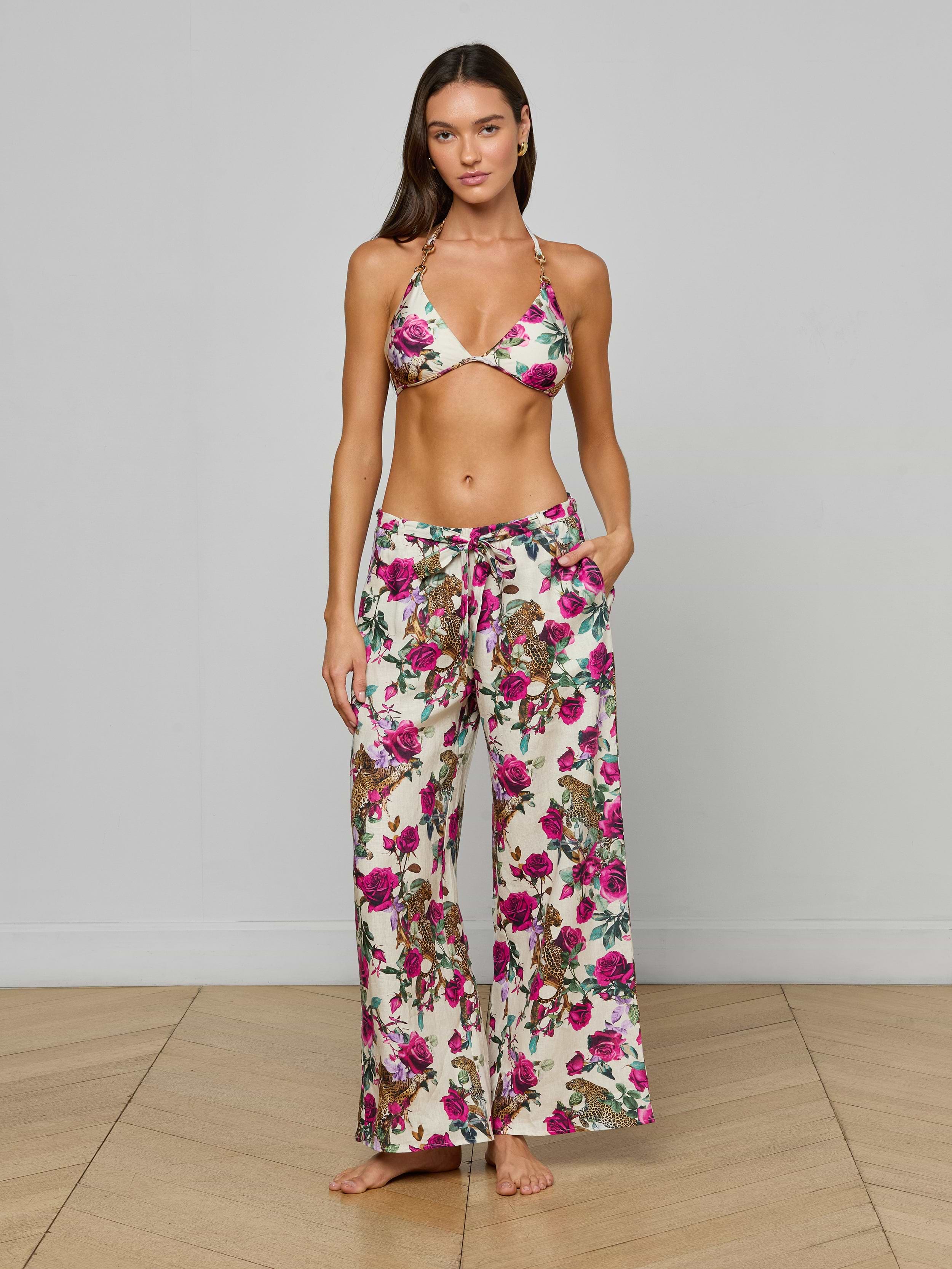 A woman stands barefoot against a plain light background, her hands tucked in her pockets, wearing the Geraldine Cover-Up Pant—a pair of loose linen resort pants with a tie waist in a rose leopard print—paired with a matching floral bikini top.