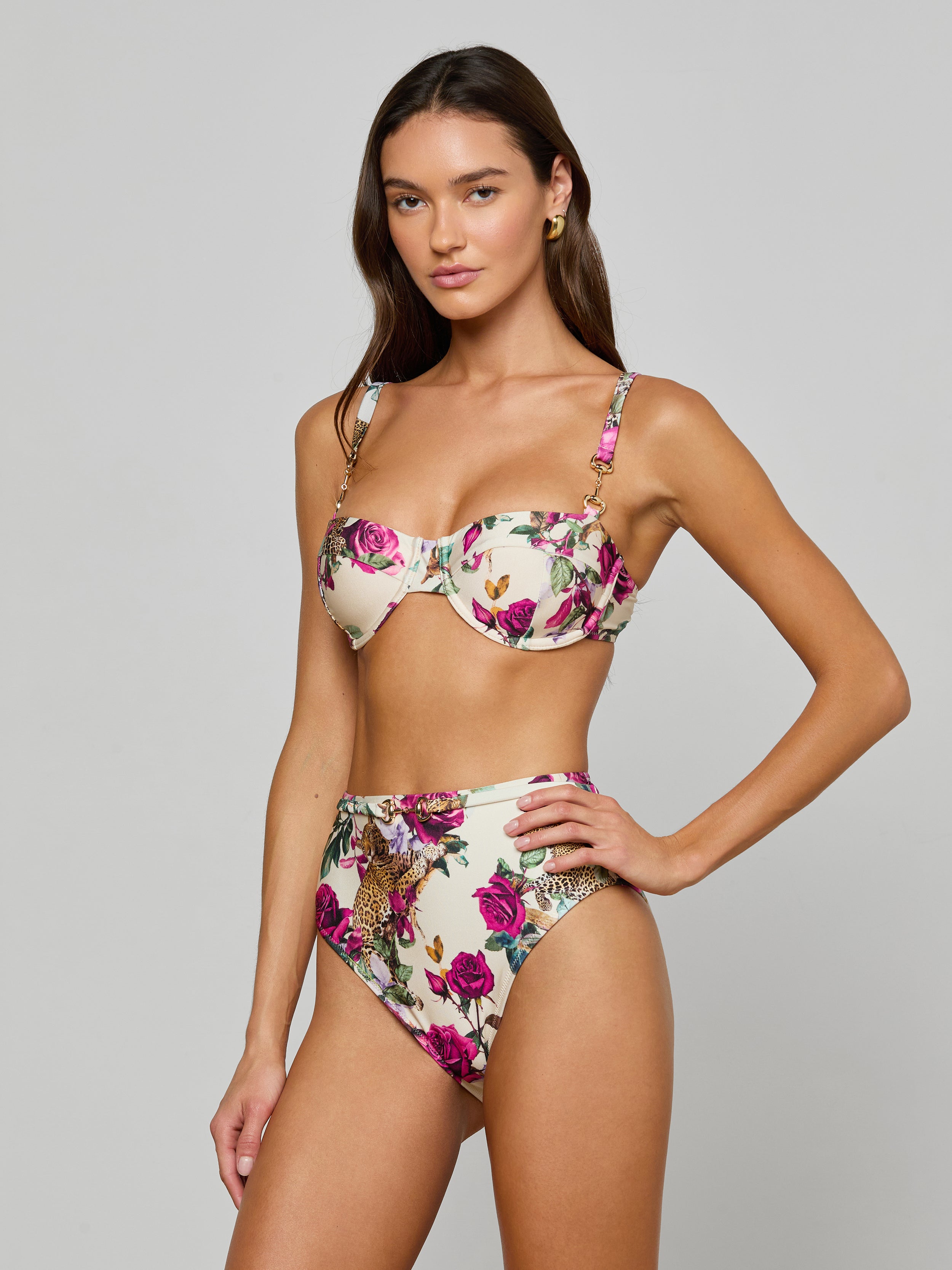 A woman poses in the Alexandria Bikini Top, a white balconette style with rose leopard print featuring underwire and adjustable straps. She pairs it with matching high-waisted bottoms, standing against a plain light background.