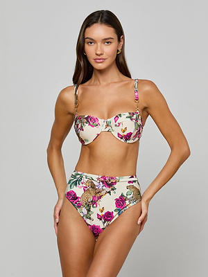 A woman with long brown hair and gold hoop earrings poses in the Vanessa High-Waisted Bikini Bottom featuring a pink, green, and beige rose leopard print. The swimwear provides UPF 50 sun protection.