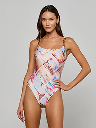 A woman models the Remi One-Piece Swimsuit with postcard print, colorful travel graphics, and luxe chain hardware straps, set against a plain light gray background.
