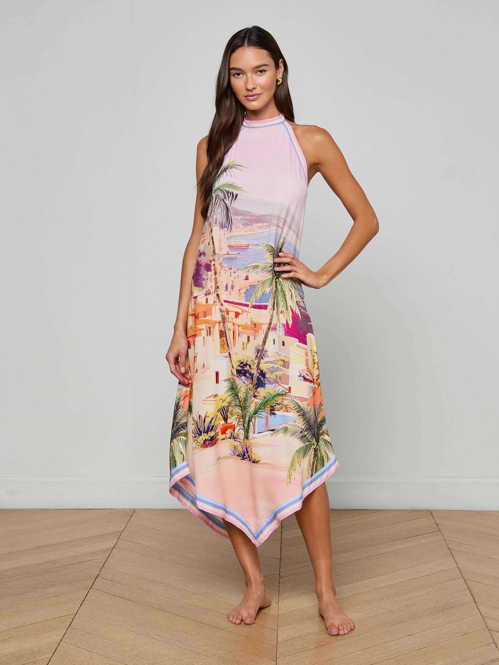 Barefoot, a woman poses against a plain white background in the Elise Cover-Up Dress—a sleeveless, halter midi dress with a handkerchief hem, featuring a vibrant scenic postcard print of a tropical city and palm trees.
