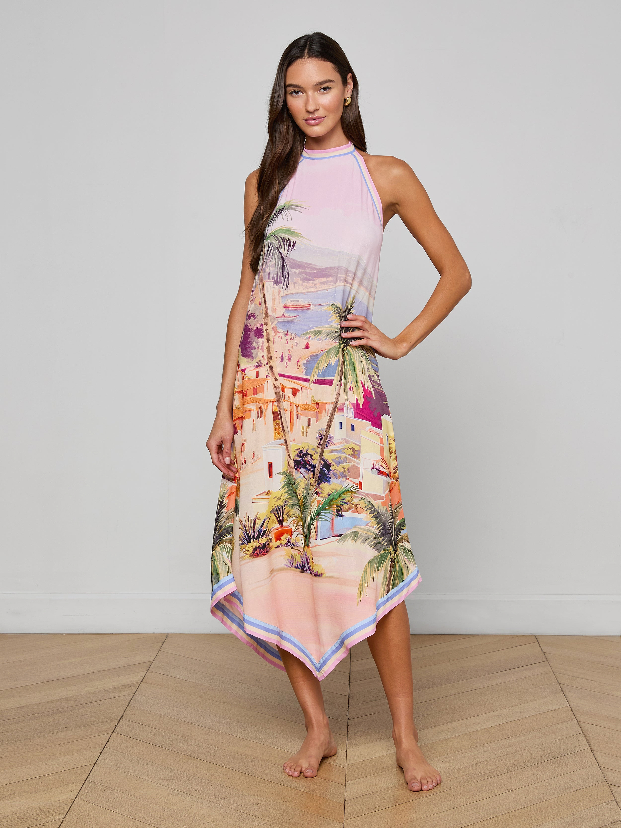 A woman stands barefoot on a wooden floor wearing the Elise Cover-Up Dress, a sleeveless pink halter midi with a scenic postcard print of a Mediterranean coastal cityscape and palm trees. She poses with one hand on her hip against a plain background.