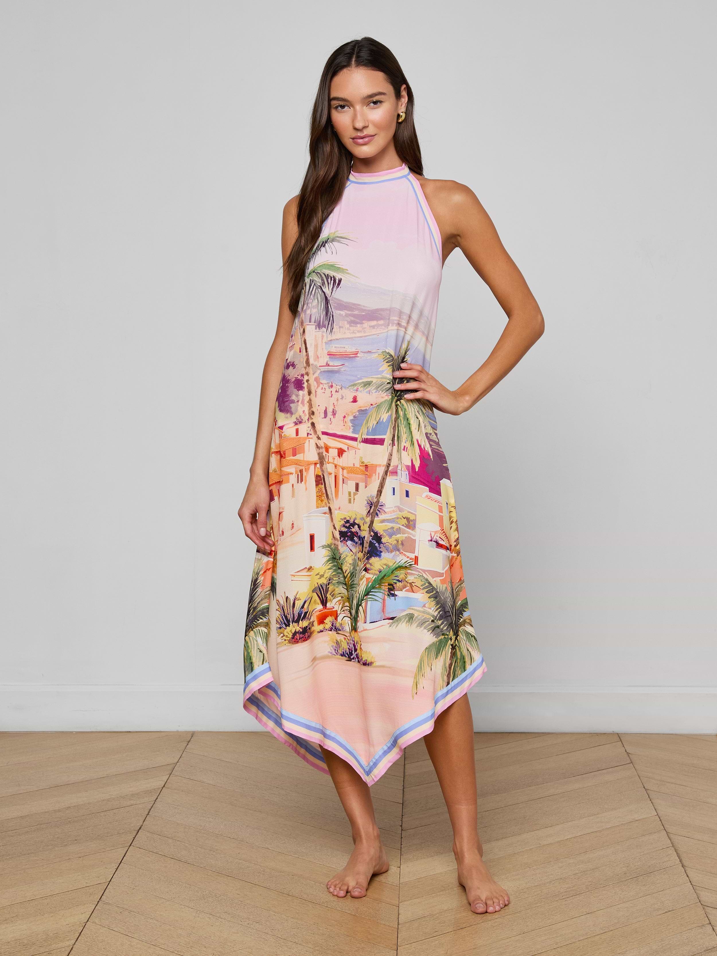A woman stands barefoot on a wooden floor wearing the Elise Cover-Up Dress, a sleeveless pink halter midi with a scenic postcard print of a Mediterranean coastal cityscape and palm trees. She poses with one hand on her hip against a plain background.