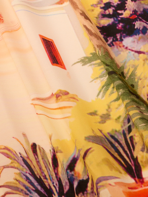 Close-up of the Elise Cover-Up Dress, featuring a vibrant scenic postcard print with green plants, architecture, a red window, and abstract patterns in yellow, green, purple, and beige.