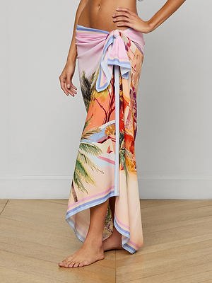 A person stands barefoot on a wooden floor, wearing the Maribel Cover-Up Pareo tied at the waist. The pareo features pastel tropical prints with palm trees, resembling a vivid postcard, as their hand rests on their hip.