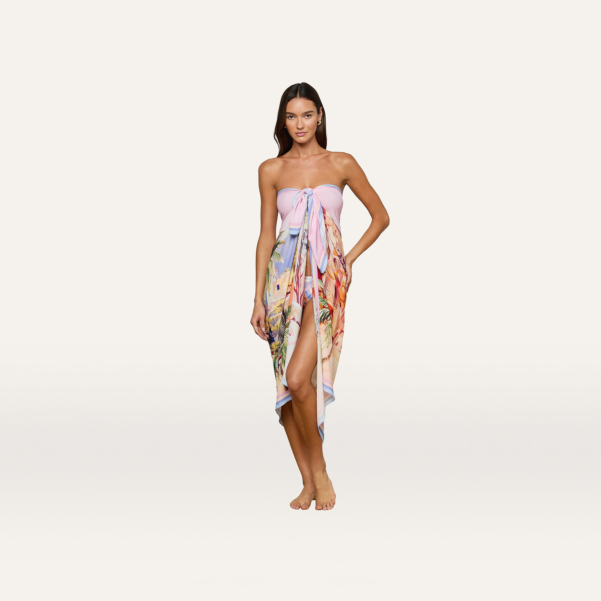 A woman stands barefoot against a plain white background, wearing the Maribel Cover-Up Pareo tied at the chest, its colorful postcard print and thigh-high slit on display.