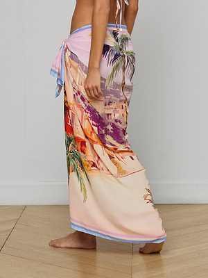 A person stands barefoot on a wood floor, wearing the Maribel Cover-Up Pareo tied at the waist, featuring a vibrant tropical and architectural print. The upper body is partially visible against a plain, light-colored background.