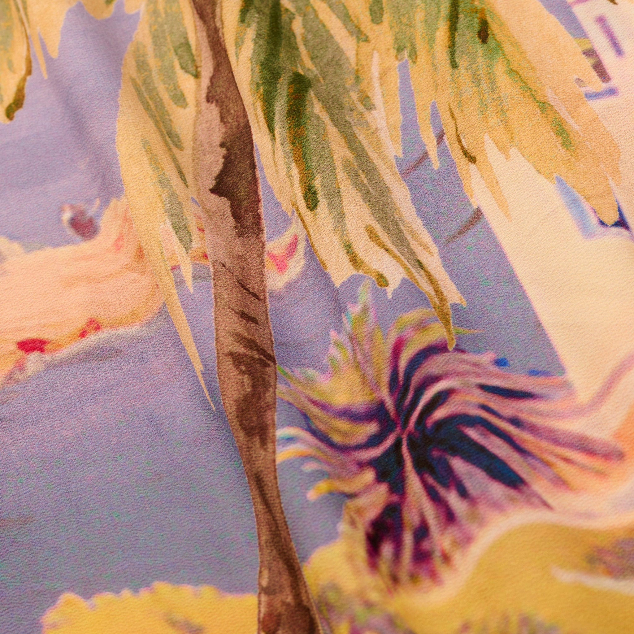 A close-up of the Maribel Cover-Up Pareo reveals its colorful, tropical postcard print, featuring palm leaves, a brown tree trunk, and blurred pastel background shapes.