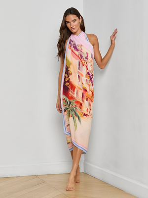 A woman stands barefoot indoors, leaning against a white wall, wearing the Maribel Cover-Up Pareo featuring a vibrant postcard print with building and palm tree motifs wrapped around her body.