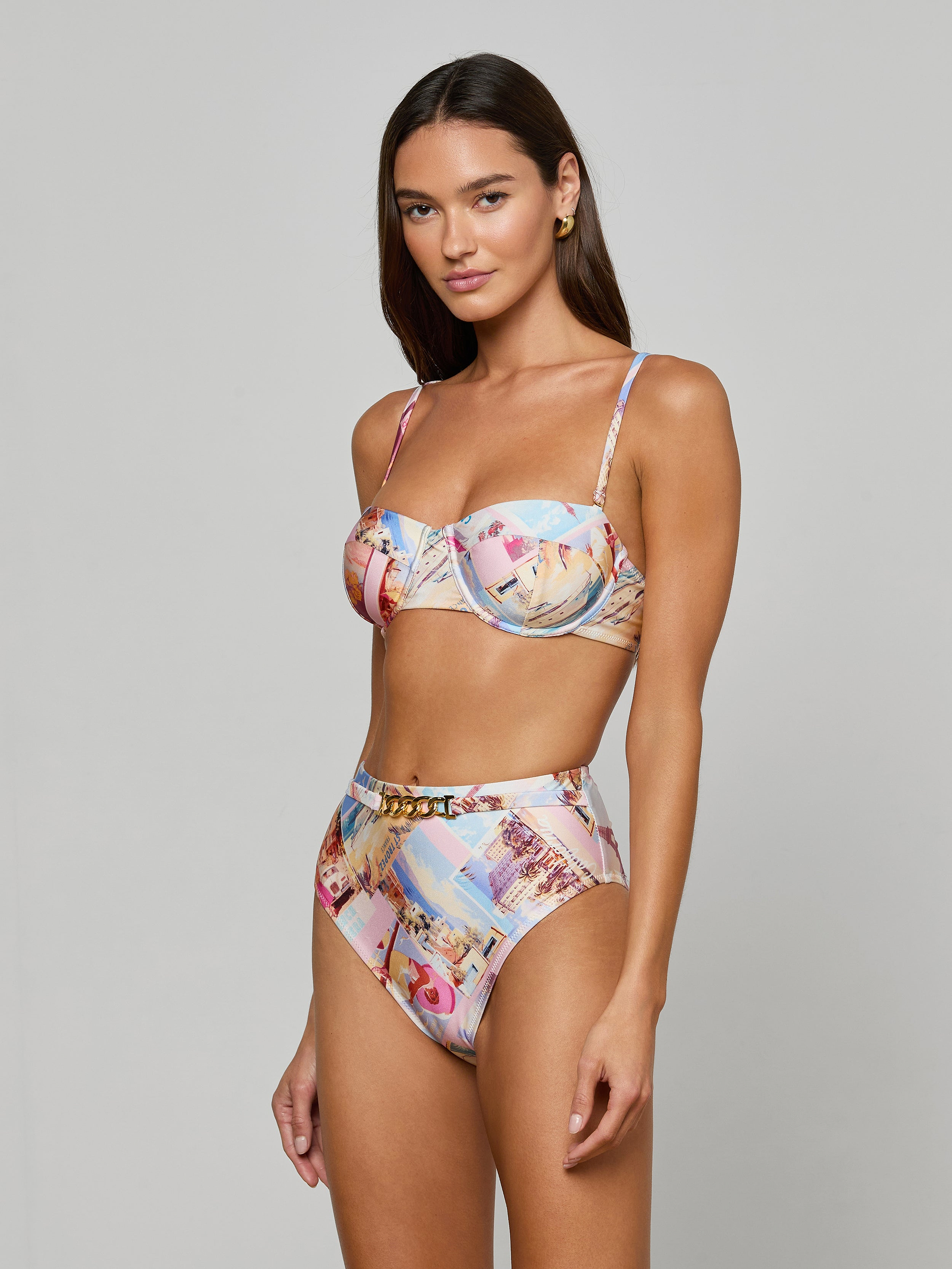 A woman wearing the Alexandria Bikini Top with high-waisted, colorful postcard print bottoms stands against a plain, light background, looking at the camera with a relaxed expression.