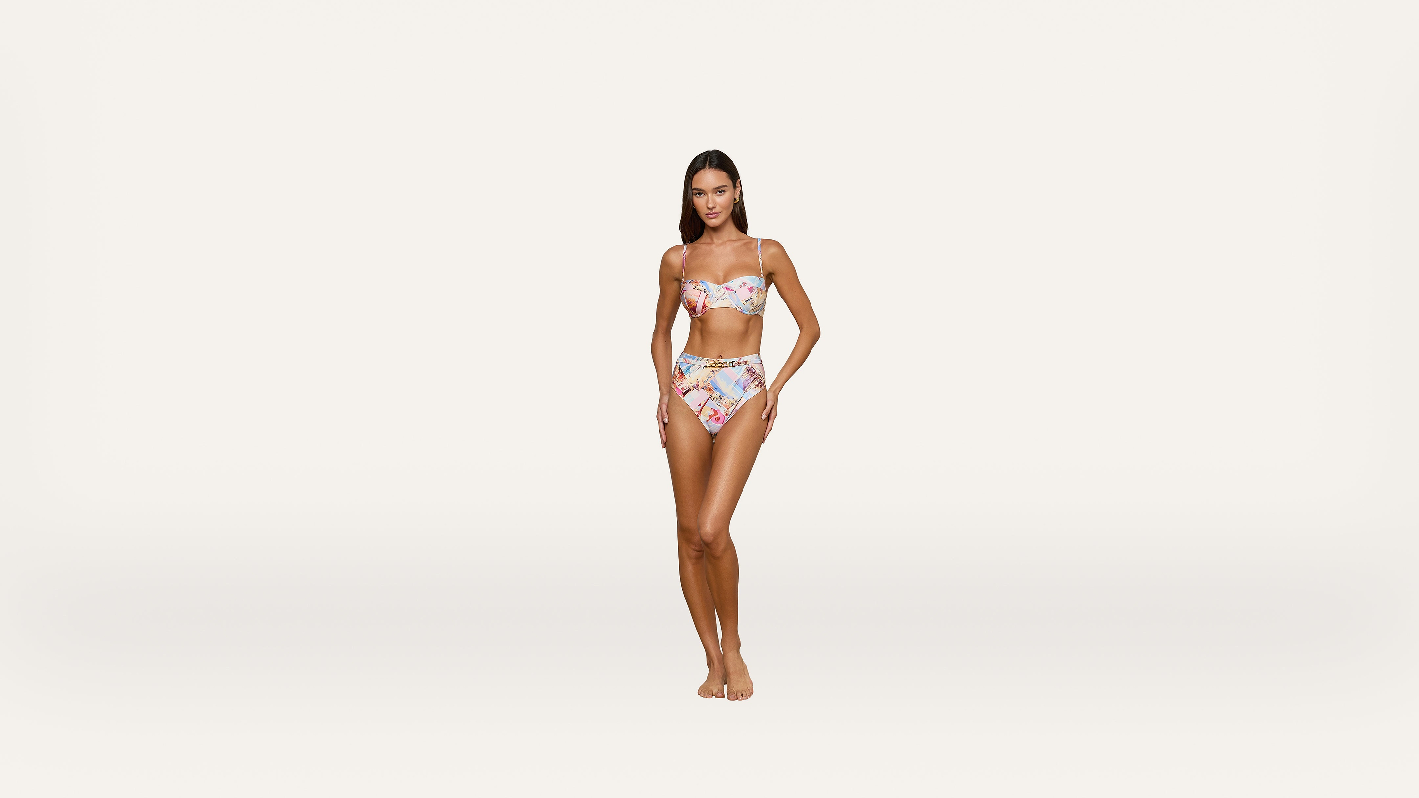 A woman stands barefoot against a plain white background, wearing a floral strapless bikini top and high-waisted bikini bottoms, with one hand resting on her hip.