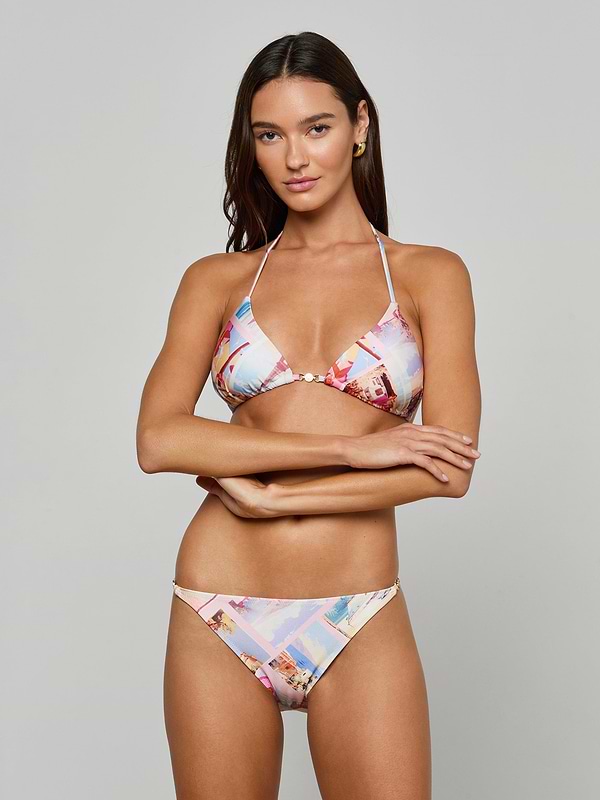 A woman with long brown hair, standing with arms crossed against a plain light background, wears the Violet String Bikini Bottom in a colorful postcard print.
