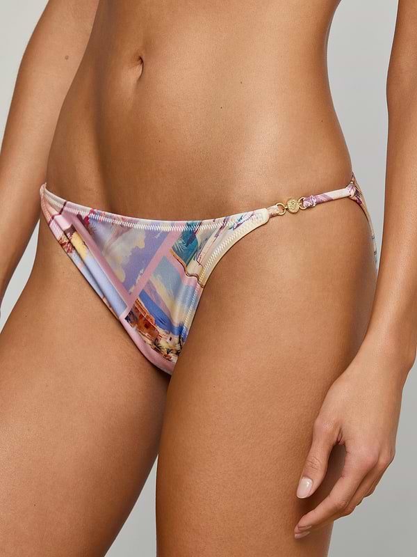 A woman models the Violet String Bikini Bottom, featuring a postcard print, thin side straps, and gold-tone circular accents. Her torso and upper thighs are visible against a plain background.