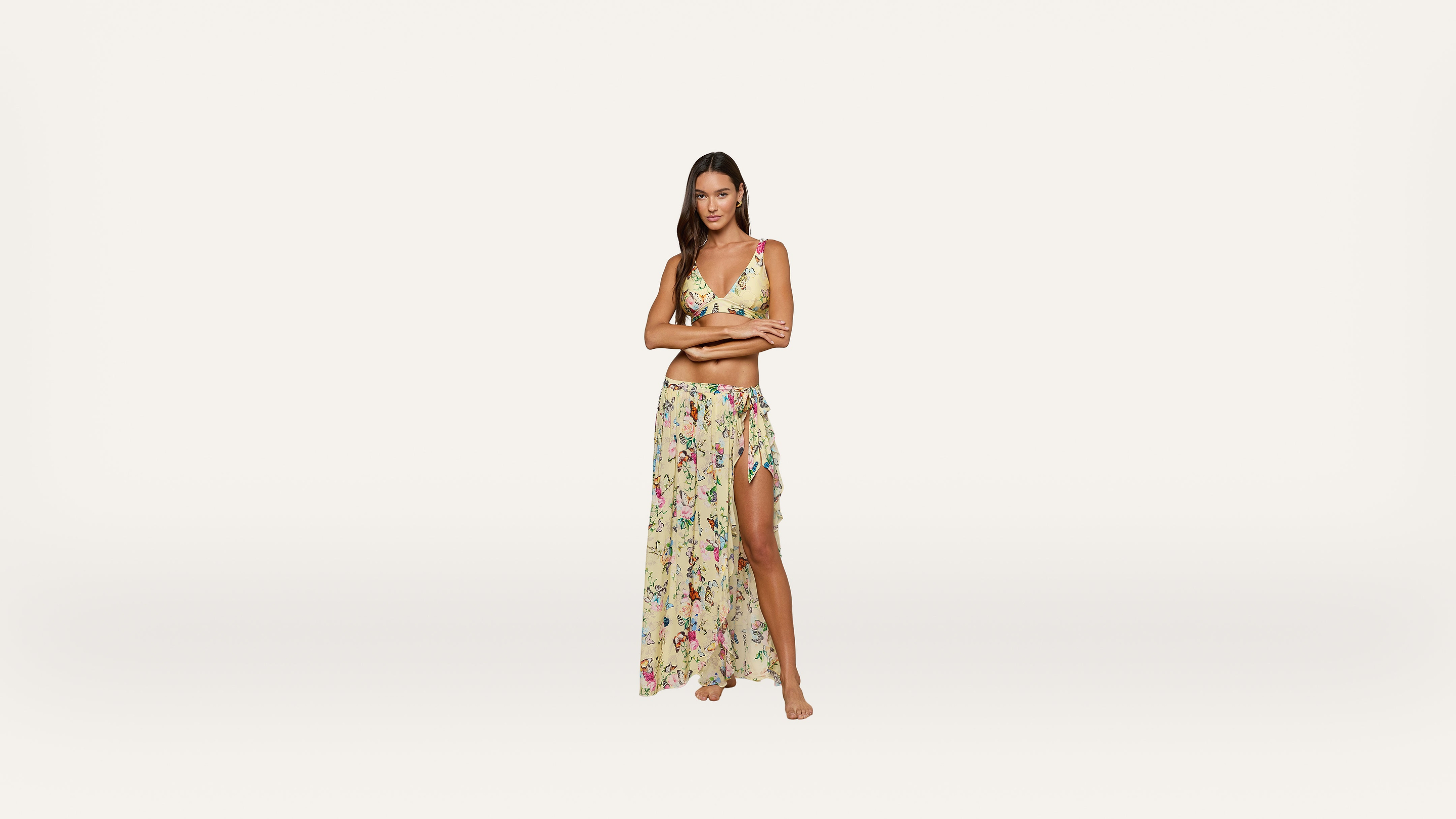 A woman stands barefoot wearing a yellow floral bikini top and a matching sheer, long skirt with a high slit, against a plain white background.
