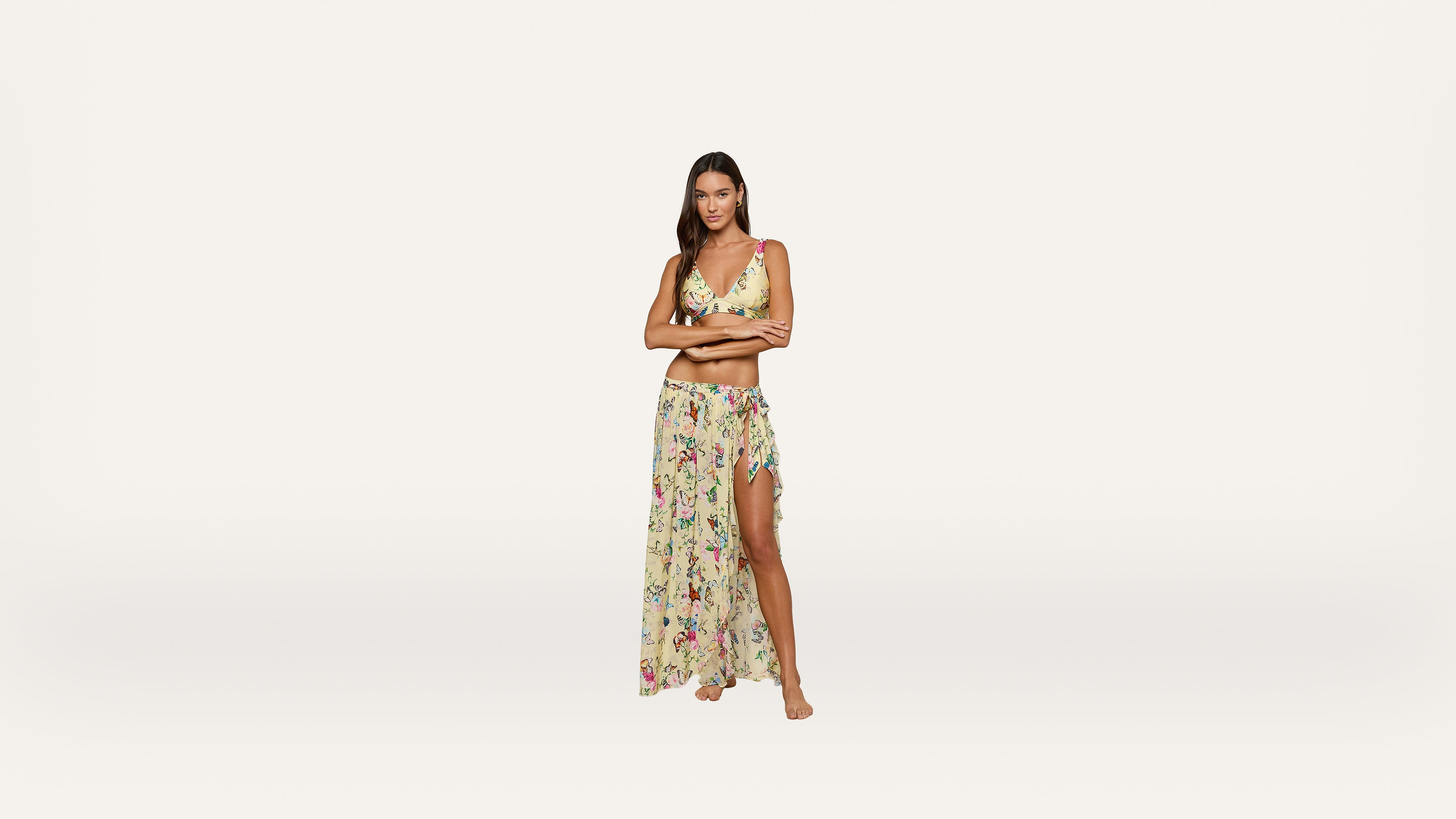 A woman stands barefoot wearing a yellow floral bikini top and a matching sheer, long skirt with a high slit, against a plain white background.