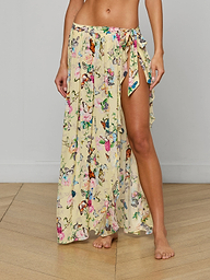 A woman stands barefoot on a wooden floor against a plain wall, wearing the Klaire Cover-Up Skirt—a long wrap maxi skirt with a butterfly print, high slit, and side tie.