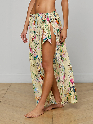 A person stands barefoot on a wooden floor, wearing the Klaire Cover-Up Skirt—a long, light yellow wrap maxi skirt with a butterfly print and high slit tied at the waist. The background is a plain, light-colored wall. Only the lower half is visible.