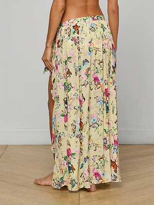 A person stands barefoot on a wooden floor wearing the Klaire Cover-Up Skirt, a long, light yellow viscose chiffon maxi with butterfly and flower patterns. The photo taken from behind highlights the skirt’s full length and vibrant butterfly print.