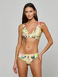 A woman poses in the Lexie Bikini Top, a yellow floral v-neck bikini featuring adjustable straps and matching bottoms, showcasing a colorful pattern against a plain light background.