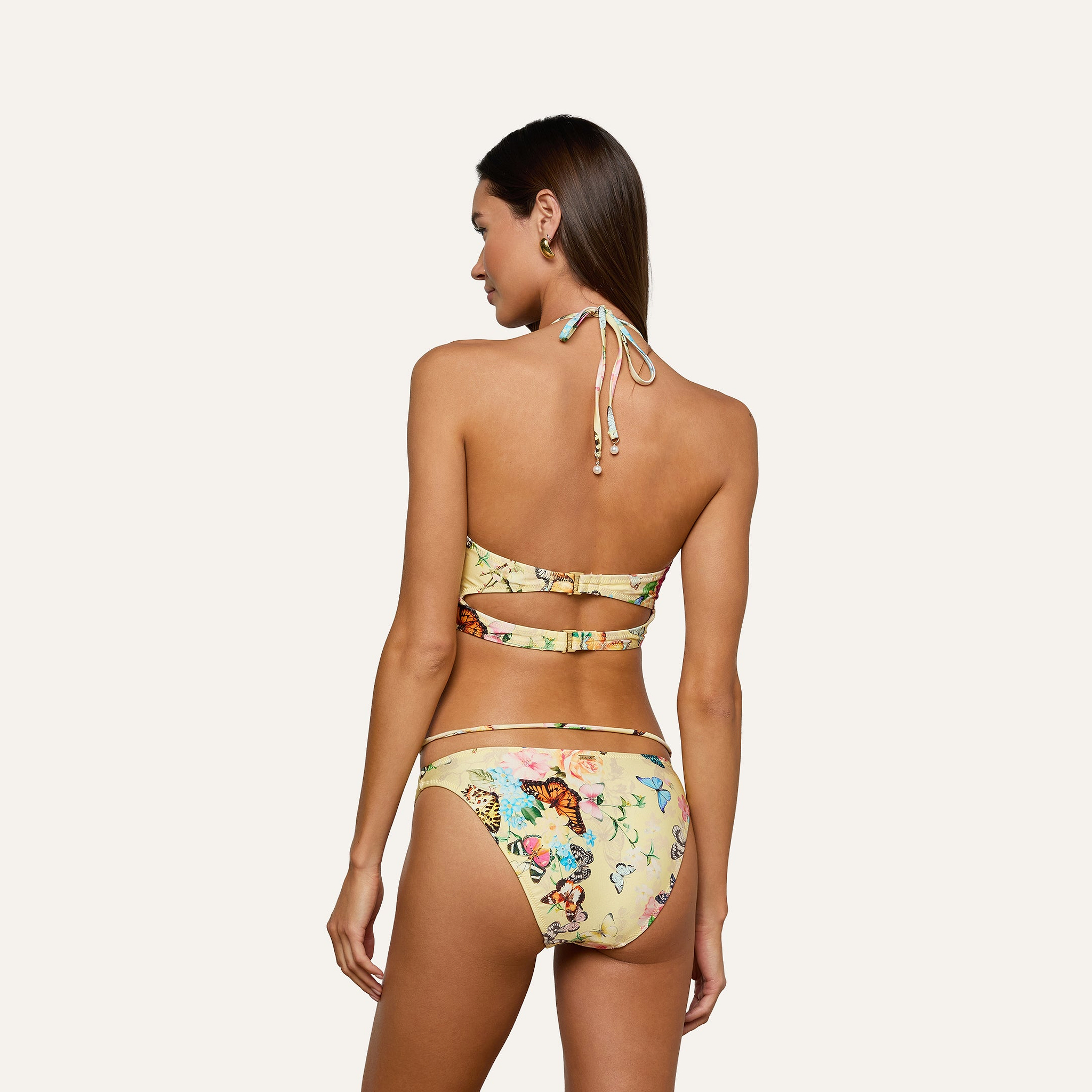 A woman with long dark hair is shown from the back, wearing the Jean Bikini Bottom featuring a butterfly print, colorful floral accents, and a high-leg cut. The yellow halter top has cut-out details. She stands against a plain light gray background.