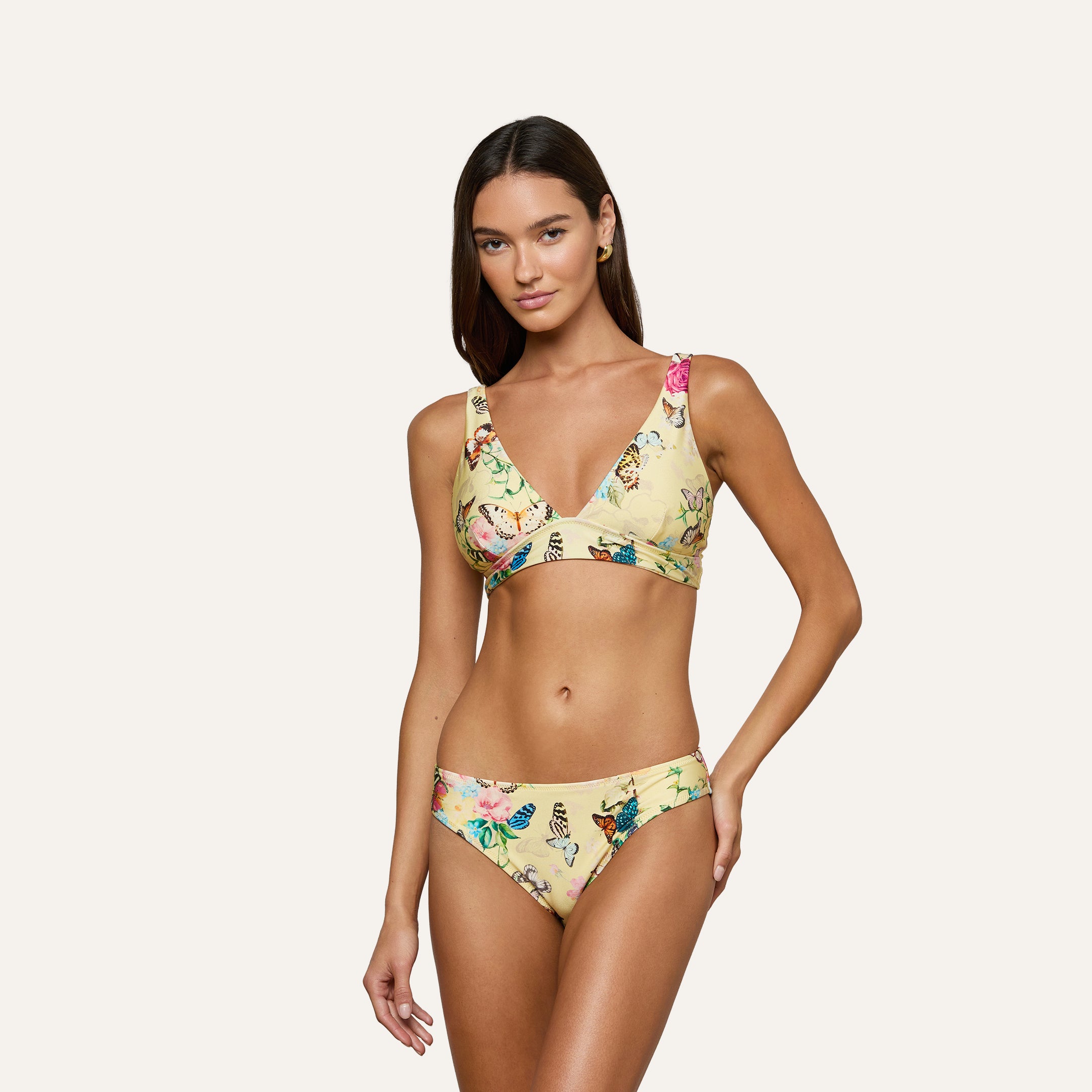 A woman poses in the Nicole Bikini Bottom, a yellow floral bikini style with moderate coverage, set against a plain light background; her left hand rests on her hip while her right arm hangs relaxed by her side.