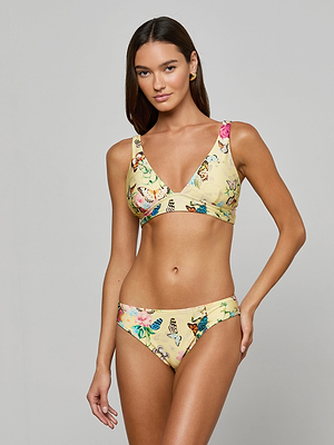 A woman with long brown hair poses against a plain background in the Nicole Bikini Bottom, featuring a colorful butterfly print on yellow UPF 50 stretch nylon fabric. She smiles slightly with one hand by her side.