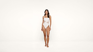 A woman stands facing forward, wearing a light pink one-piece swimsuit with cut-out lace details on the sides, against a plain white background.