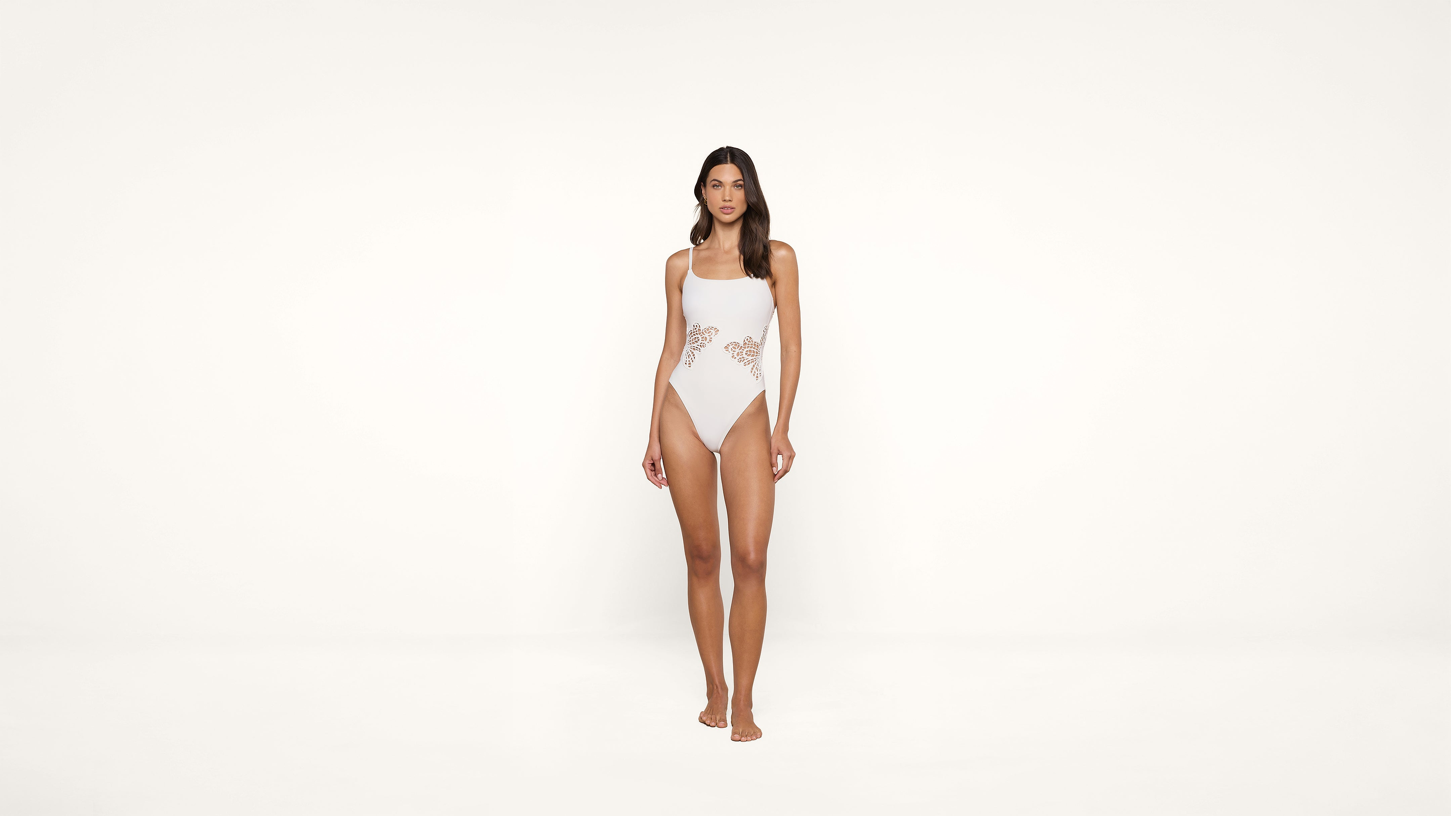 A woman stands facing forward, wearing a light pink one-piece swimsuit with cut-out lace details on the sides, against a plain white background.