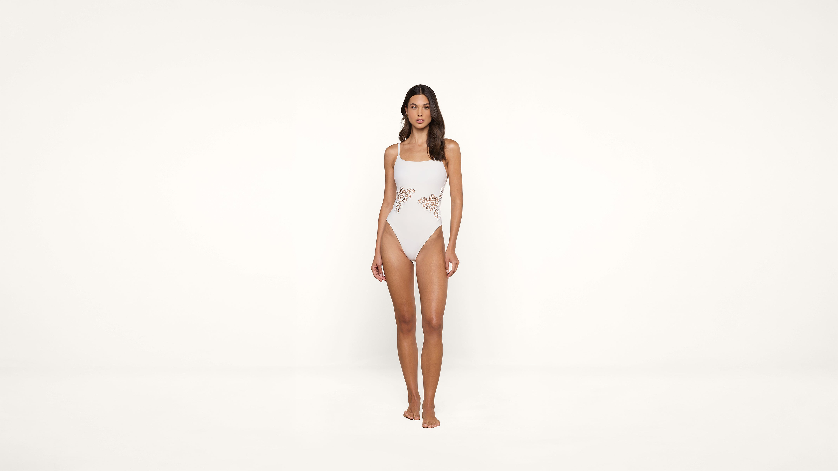 A woman stands facing forward, wearing a light pink one-piece swimsuit with cut-out lace details on the sides, against a plain white background.