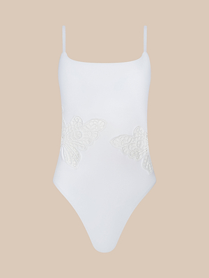 The Remi One-Piece Swimsuit features thin straps, a high-leg cut, and delicate semi-transparent butterfly lace appliqué on the sides, shown against a beige background.