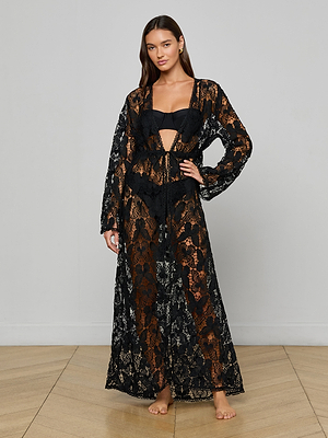 A woman stands barefoot indoors, gracefully posing against a light gray wall and wooden floor in the Janus Cover-Up Robe—a sheer black bohemian piece with a floral pattern and long sleeves, styled over her bikini.