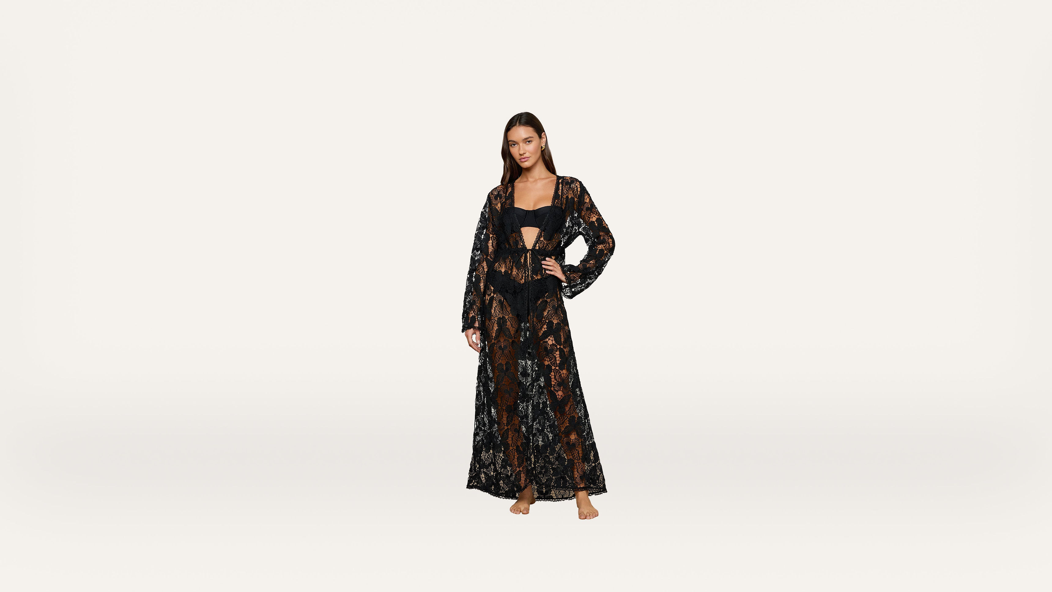 A woman stands barefoot, wearing a long, sheer black lace dress over a matching black bralette and high-waisted shorts, posing against a plain white background.