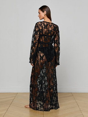 A woman stands barefoot on a wooden floor, facing away from the camera, wearing the Janus Cover-Up Robe— a long, sheer black lace bohemian robe with long sleeves over a black undergarment set. The background is a plain white wall.