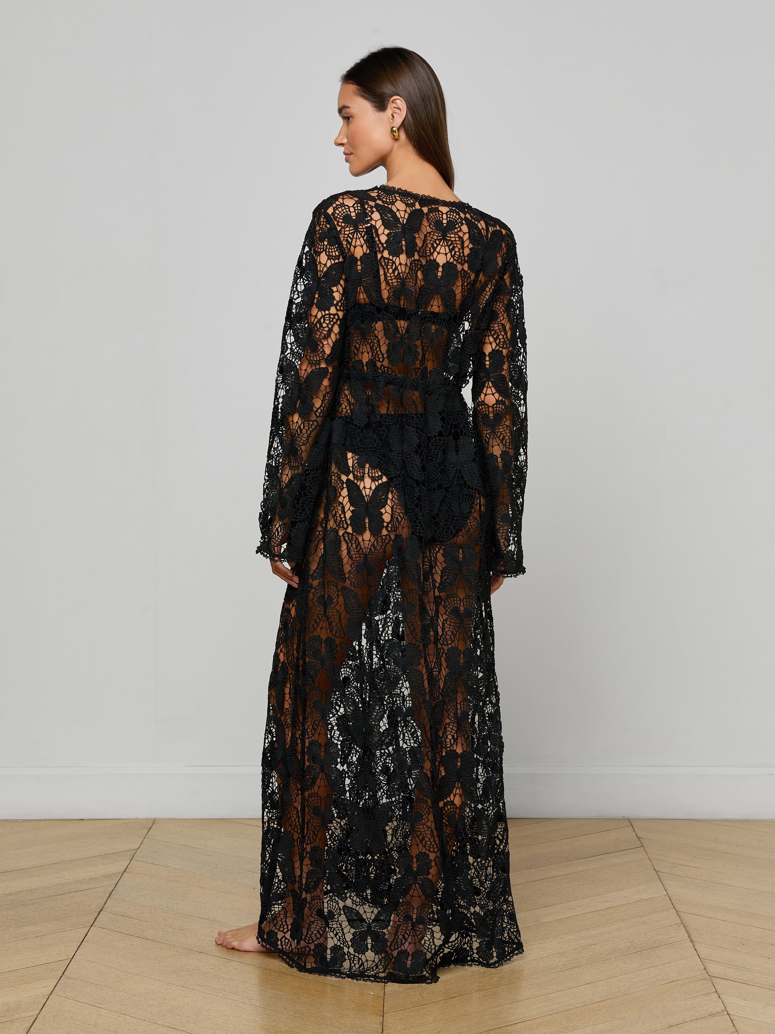 A woman stands barefoot, facing away, wearing the Janus Cover-Up Robe—a long, sheer black lace maxi beach dress with long sleeves. The robe features a floral pattern and reveals her silhouette against a plain white background.