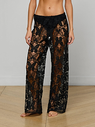 A person stands barefoot against a plain white background, wearing the Geraldine Cover-Up Pant. These black, see-through butterfly crochet lace pants feature a brown underlayer, a loose wide-leg fit, and a low waist—perfect for effortless bohemian resort wear style.