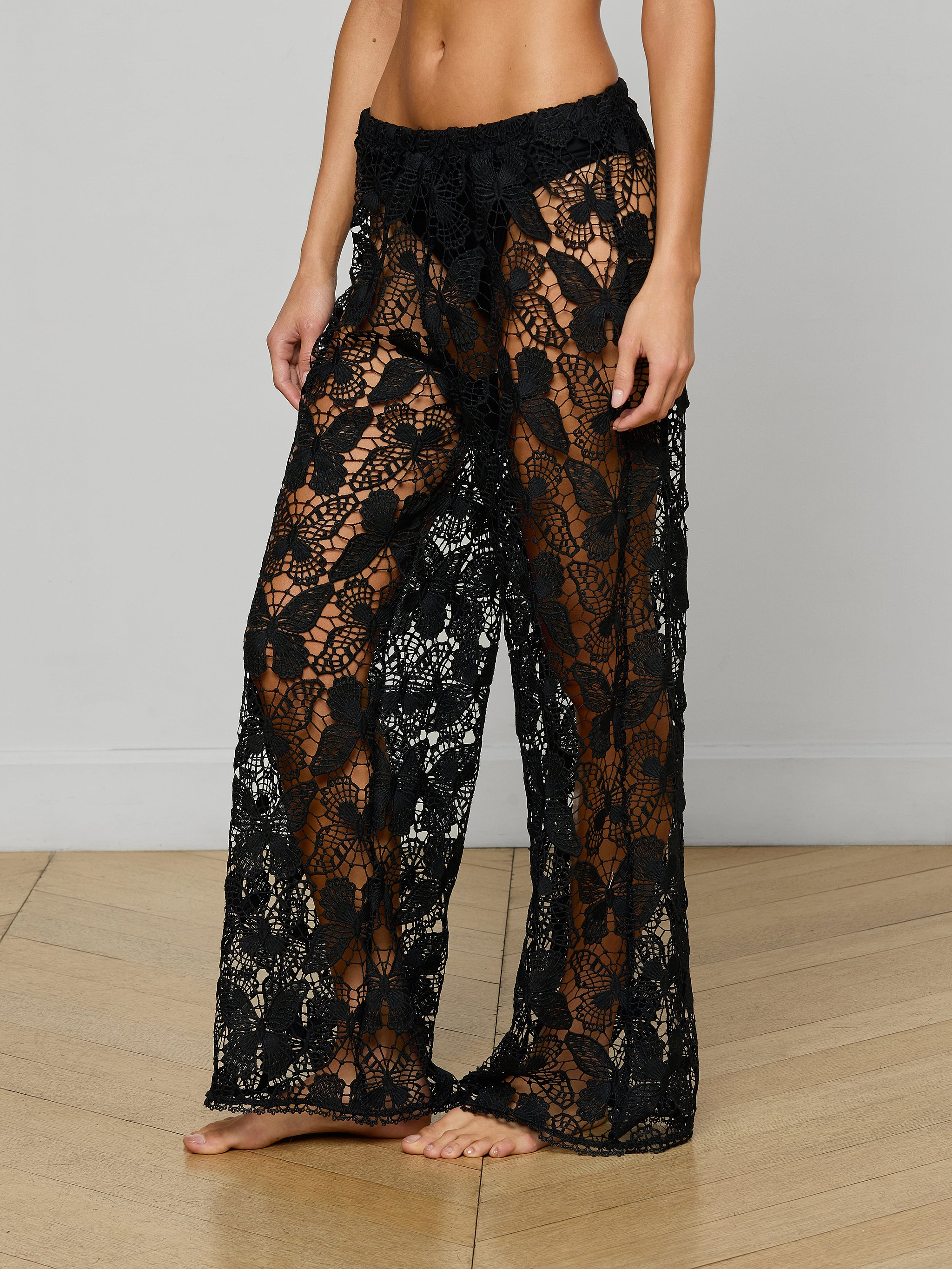 Model wearing the Geraldine Cover-Up Pant—black, sheer, lace wide-leg pants featuring a floral pattern and bohemian flair—standing barefoot against a plain white background.
