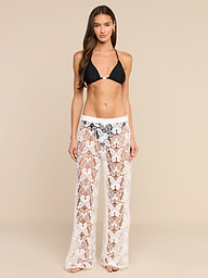 A woman stands against a plain background, facing forward with a neutral expression, wearing a black bikini top paired with the Geraldine Cover-Up Pant—white sheer lace bohemian pants.