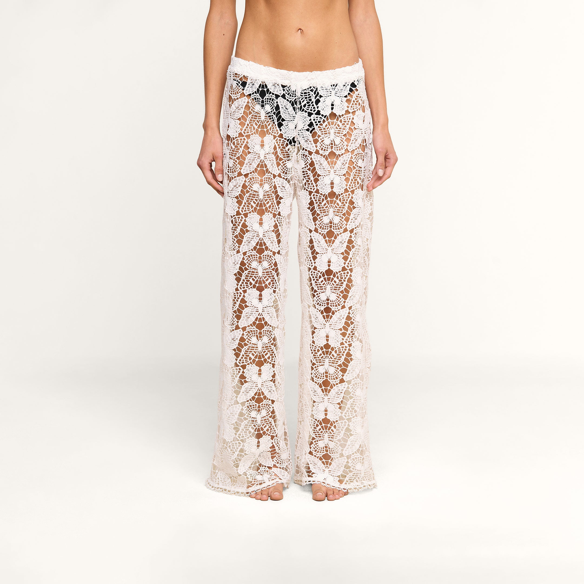 A person wearing the Geraldine Cover-Up Pant—sheer, wide-leg, white crochet lace with a floral pattern—stands barefoot against a plain, light background.