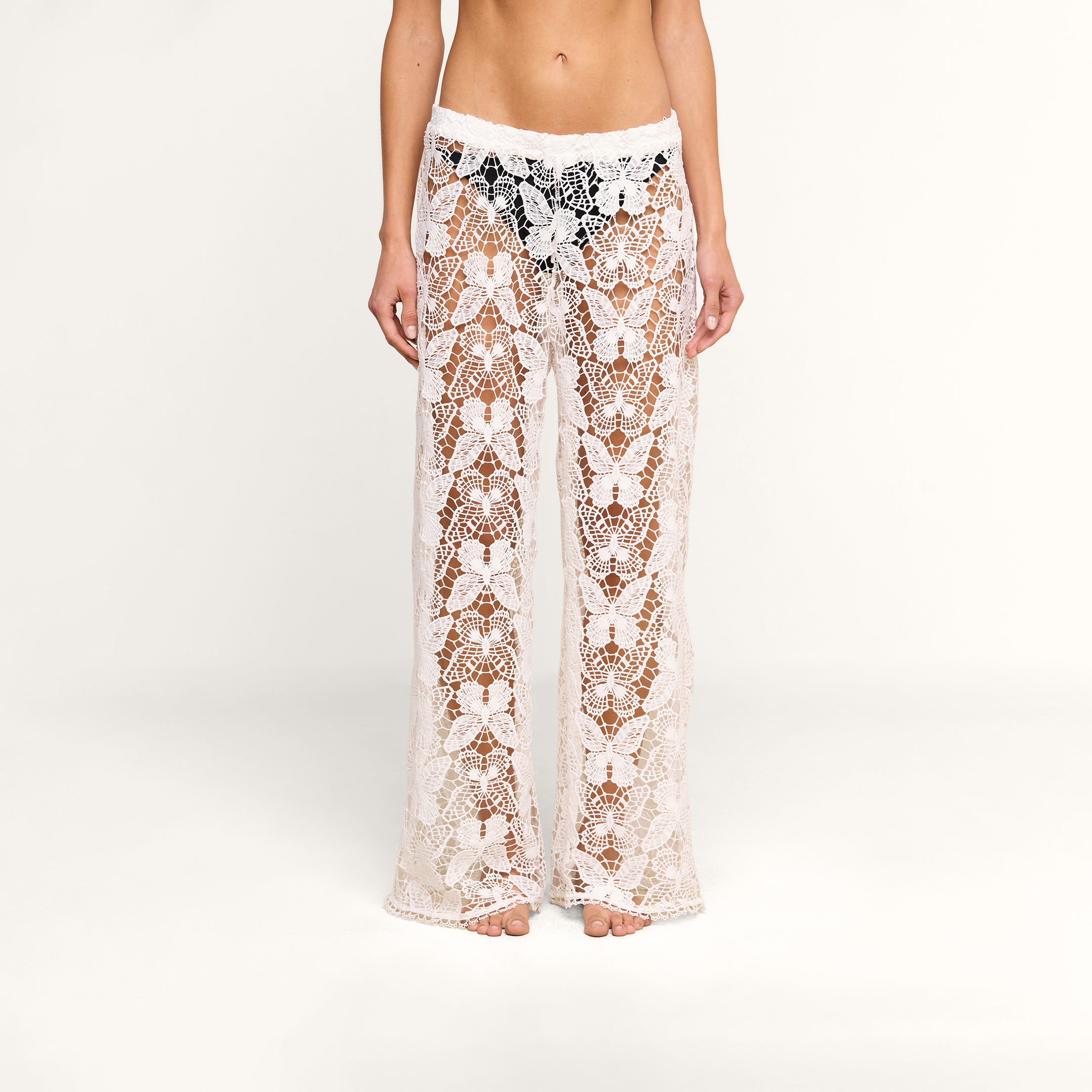 A person wearing the Geraldine Cover-Up Pant—sheer, wide-leg, white crochet lace with a floral pattern—stands barefoot against a plain, light background.