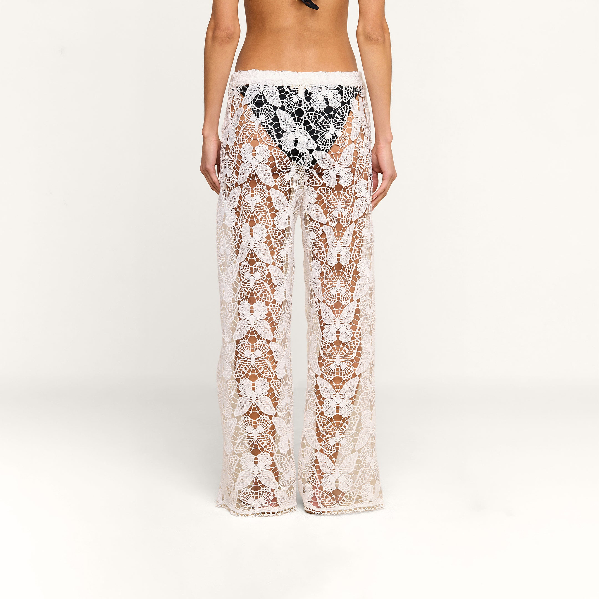 A person is shown from the back against a plain white background, wearing the Geraldine Cover-Up Pant—sheer, white lace bohemian pants—over black underwear.
