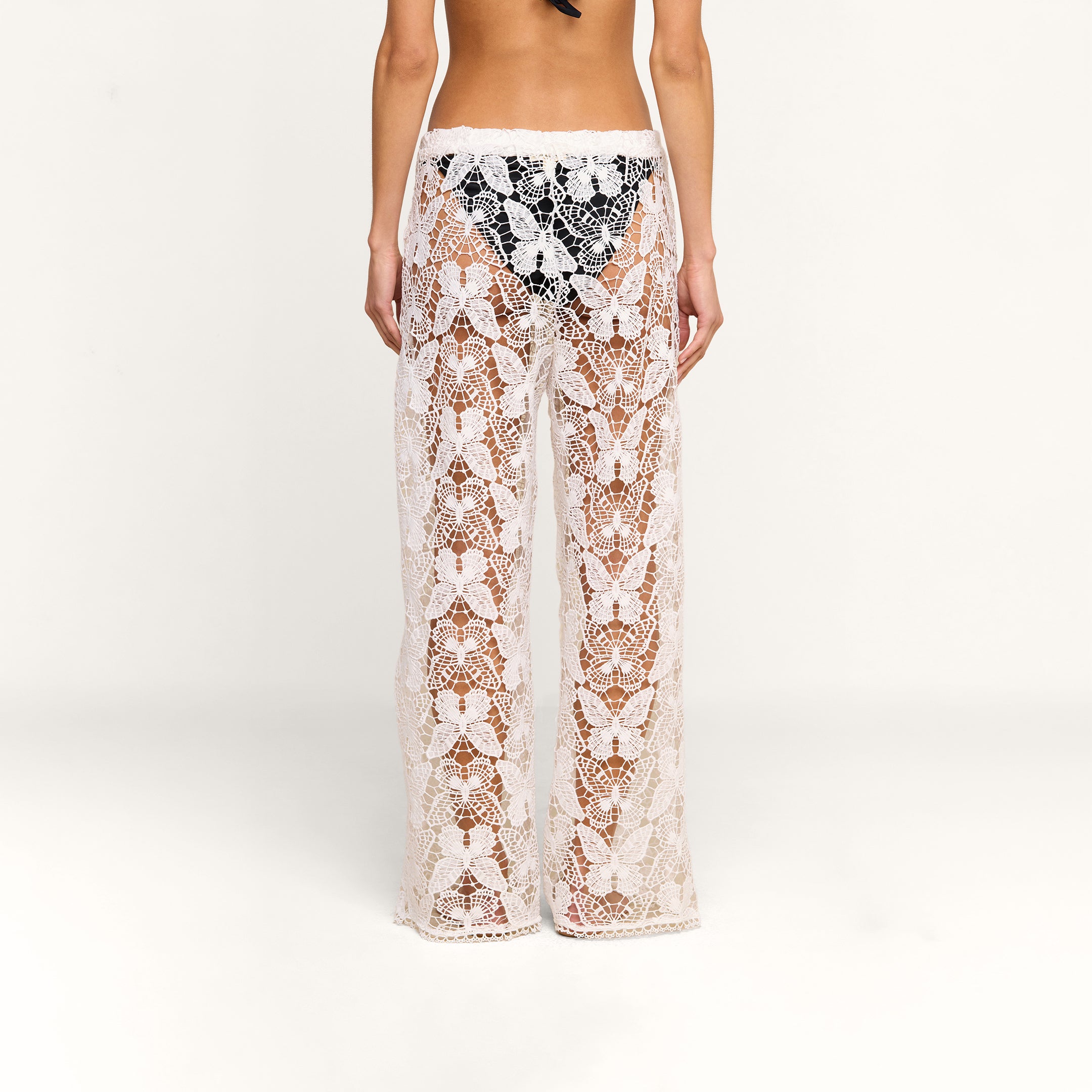A person is shown from the back against a plain white background, wearing the Geraldine Cover-Up Pant—sheer, white lace bohemian pants—over black underwear.