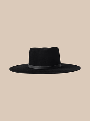 Il cappello Fedora con fascia in pelle Reyes è un cappello nero realizzato a mano, caratterizzato da una tesa larga, una corona piatta e una sottile fascia in pelle, raffigurato su uno sfondo beige.