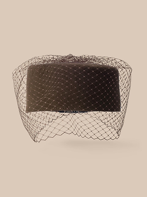 El sombrero «Anstine Pillbox» en color negro, adornado con un delicado velo tipo jaula, se presenta sobre un fondo beige y irradia sofisticación vintage.