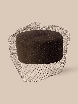 Der „Anstine Pillbox“-Hut strahlt mit seinem schwarzen Design und dem zarten Vogelkäfig-Schleier Vintage-Eleganz aus; er wurde vor einem schlichten beigen Hintergrund fotografiert.