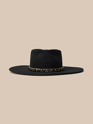 The Ferrante Hat is a handcrafted, wide-brimmed black felt hat made in LA, featuring a gold chain band around the base of the crown, displayed against a beige background.