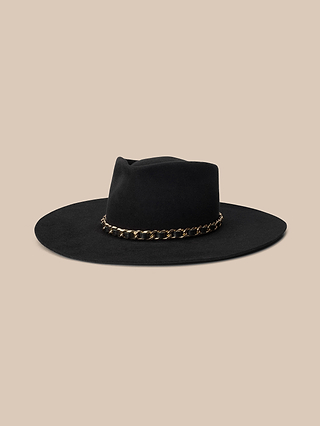 The Ferrante Hat—a black, wide-brimmed felt hat handcrafted in LA—features a gold chain band and is displayed against a beige background.