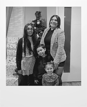 A woman and three children stand smiling in front of a decorated Christmas tree and wrapped gifts inside a home. The image is in black and white.